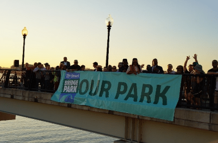 A group of people stands on a bridge holding a banner that reads "OUR PARK" at sunset.