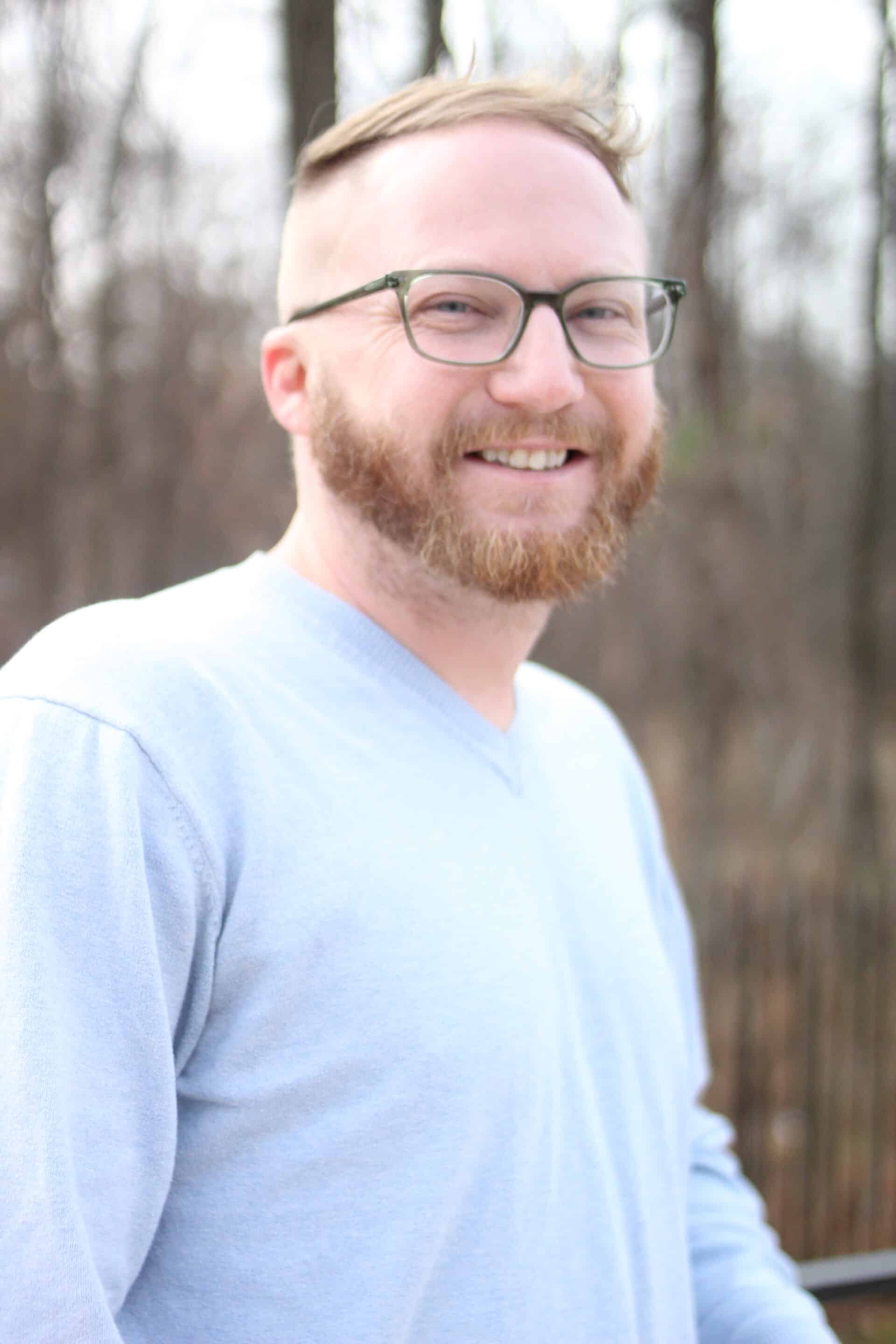 A bearded person from the 11th Street Bridge Park Team, wearing glasses and a light blue shirt, smiles outdoors with blurred trees in the background, embodying the spirit of building a better future by bridging communities.