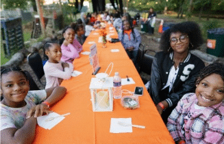 A group of people seated at a long table covered with an orange tablecloth, outdoors, with trees in the background.