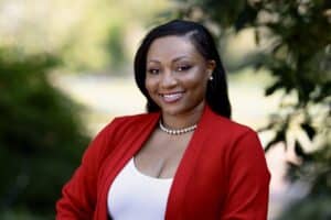 A woman in a red blazer and white top stands outdoors, smiling as part of the 11th Street Bridge Park Team - Bridging Communities, dedicated to building a better future.