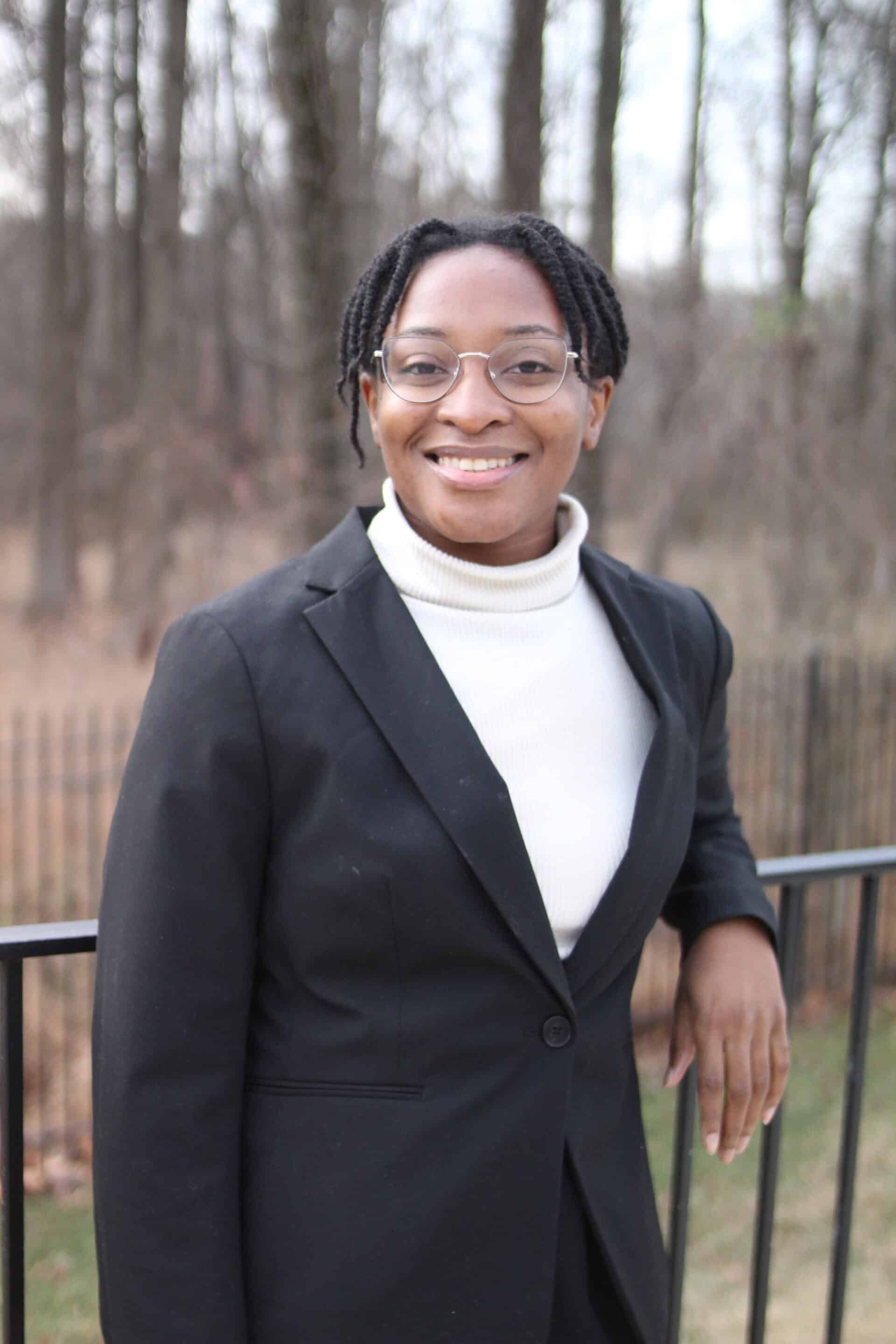 A member of the 11th Street Bridge Park Team, wearing glasses and a black blazer over a white top, stands by a fence with trees in the background, embodying the spirit of bridging communities and building a better future.