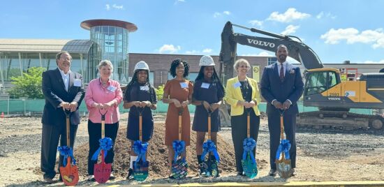 Seven people in business attire and hard hats stand with shovels at a construction site, embodying the spirit of "Building Bridges Across The River." A Volvo excavator is poised in the background, ready to transform ideas into reality.