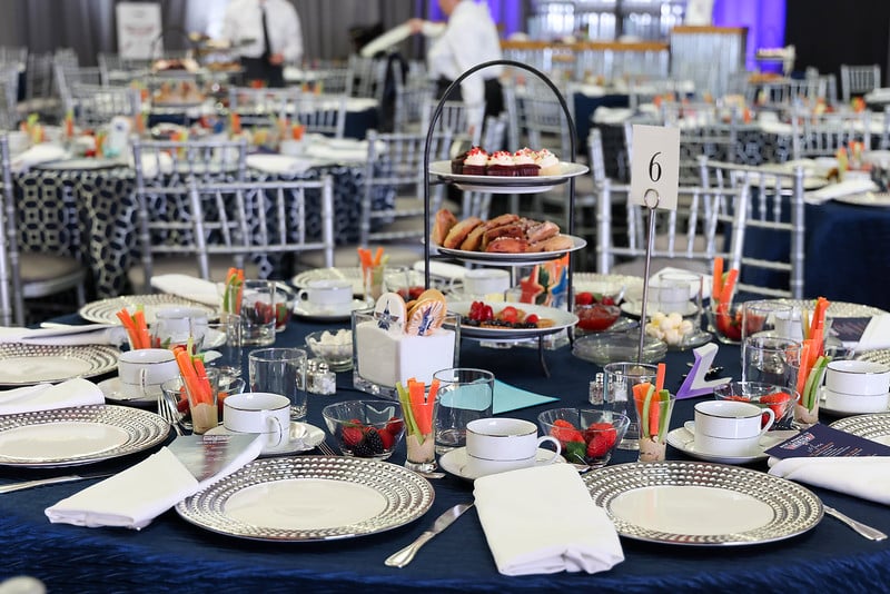 The Annual Wacky and Whimsical Washingtonian Tea presents a set dining table adorned with tiered trays of pastries, plates, utensils, glasses, and small bowls of fruit. A dark blue tablecloth drapes elegantly over the setting, surrounded by silver chairs.