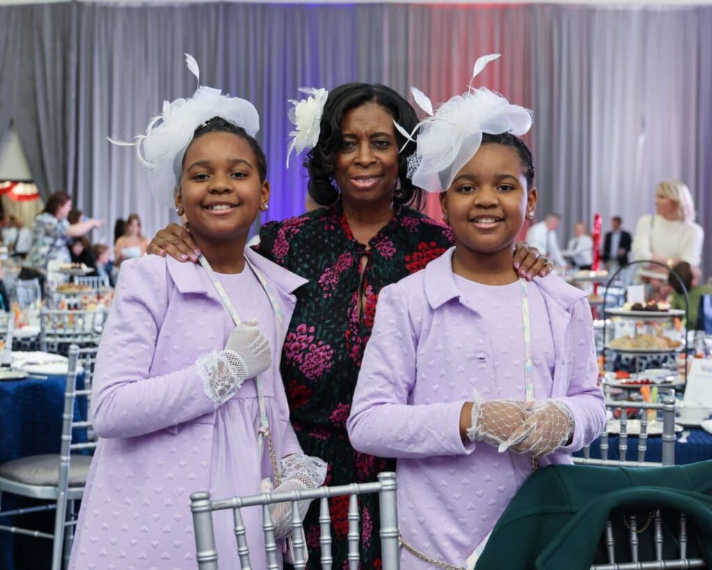 At the AfterDark@THEARC Gala, three people are smiling at a formal event. Two young girls in lavender dresses stand with an older woman in a floral dress, capturing the elegance of the evening.