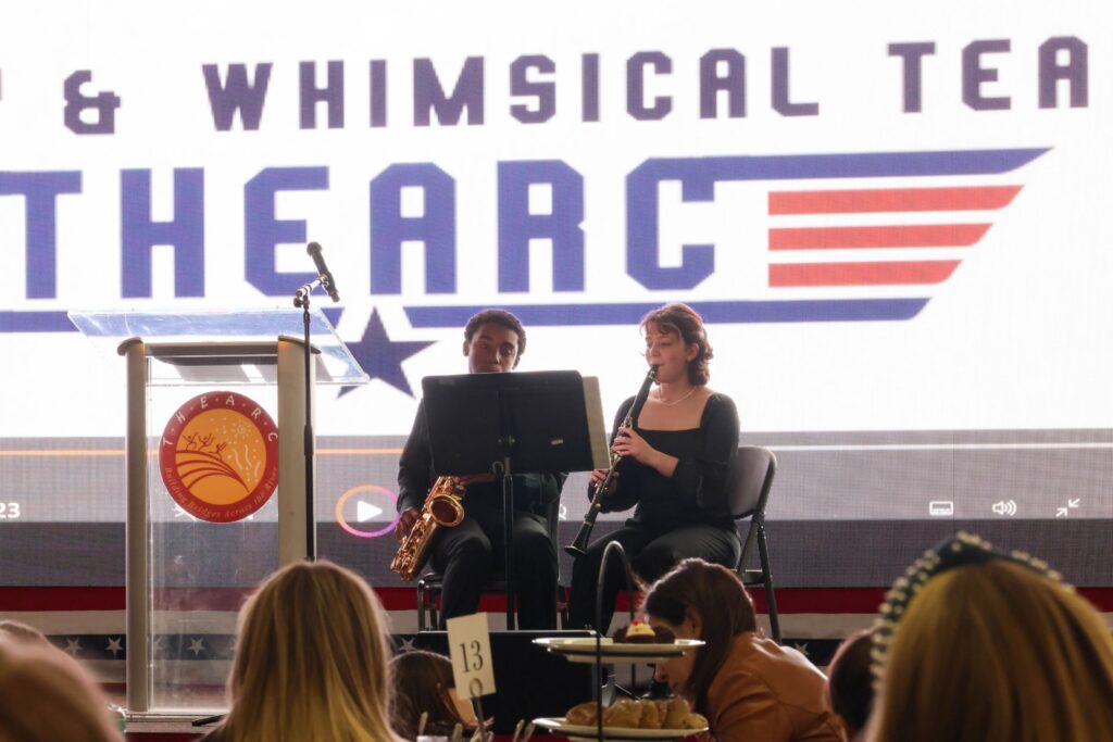 Two musicians seated on chairs, one playing a saxophone and the other a clarinet, perform on stage with a podium and large screen displaying the words "Annual Wacky and Whimsical Washingtonian Tea" in the background.