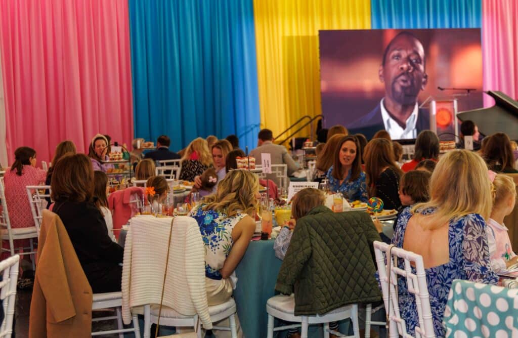 Guests at the Gala, seated at tables in a vibrant event space adorned with pink, yellow, and blue curtains, attentively watch the large screen where a man speaks. The air buzzes with excitement as AfterDark@THEARC brings the evening to life.