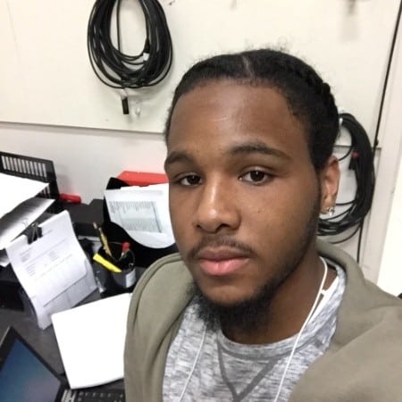 In an office setting, a person with braided hair is near a desk scattered with papers, cables on the wall, and office supplies, embodying the spirit of building bridges towards a better future.