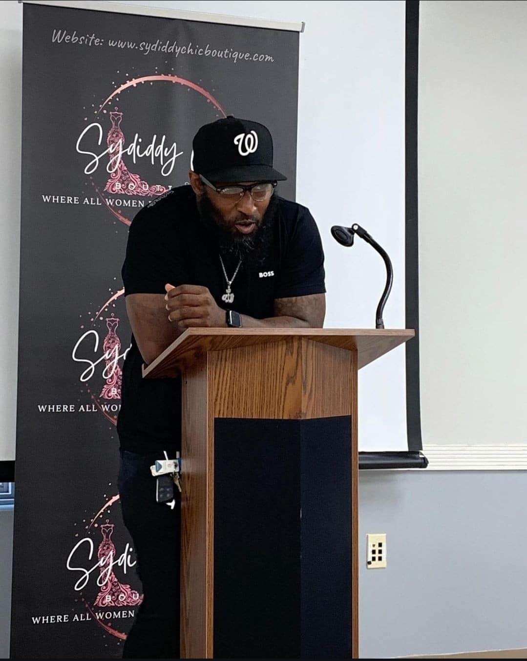 A person wearing a black cap and t-shirt stands at a podium, passionately speaking about building a better future. A "Sydiddy" banner waves proudly in the background, symbolizing the importance of building bridges within the community.