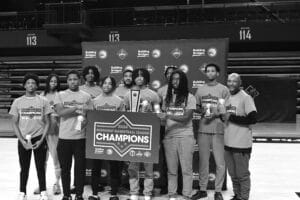 A basketball team poses with a "2024 Midnight Basketball League Champions" banner and trophies in a gymnasium.