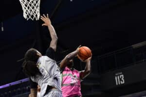 Two basketball players in action near the hoop; one wearing a gray jersey defends while the other in a pink jersey attempts a shot.