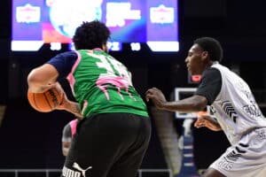 Two basketball players in action on a court, one wearing a green and pink jersey holding the ball, the other in a white and black jersey defending.