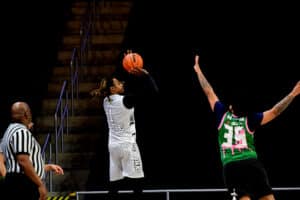 Basketball player in white jersey taking a jump shot while defender in green jersey attempts to block. Referee watches from the side.