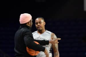 Two men are on a basketball court; one is wearing a black shirt and pink hat holding a basketball, and the other is in a white jersey, engaged in conversation.