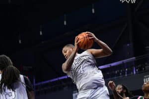 Basketball player in gray jersey holding the ball, surrounded by opposing players on a court.