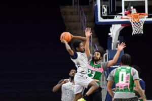 A basketball player in a white and blue uniform jumps to shoot, evading defenders in green and pink uniforms, near the hoop during a game.