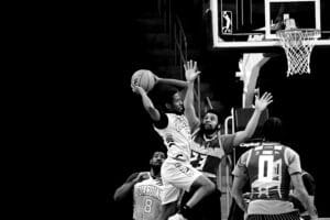 A player in a white jersey leaps towards the basketball hoop, holding the ball, while a defender in a striped jersey attempts to block. Other players watch the action.