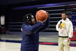 A person in a blue jacket aims to shoot a basketball in a gym while another person holds a microphone nearby.