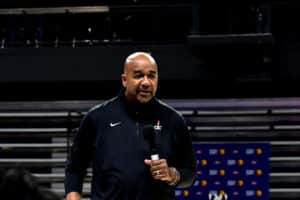 A man in a black Nike jacket holding a microphone stands in front of bleachers in an indoor arena.