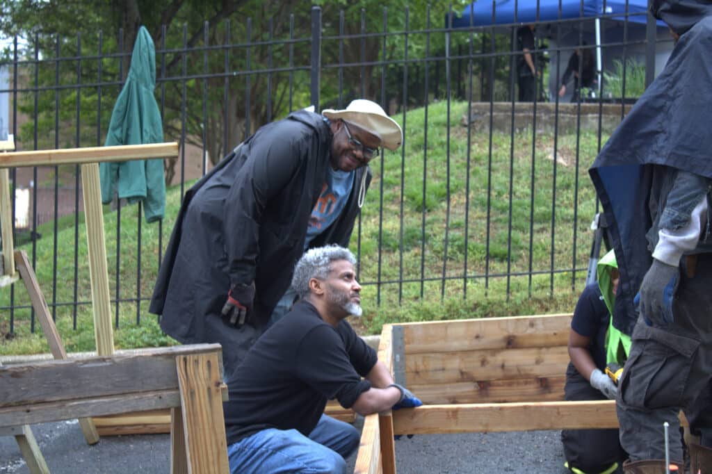 Two men work outdoors assembling a wooden structure near a fence; one man kneels, while the other stands and smiles, wearing a hat and raincoat.