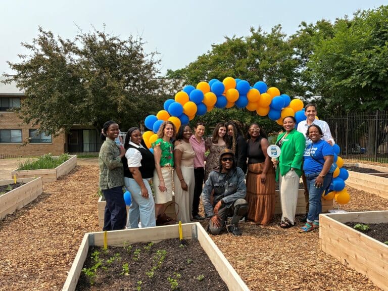 A group of people pose together in a community garden under an arch of blue and yellow balloons, surrounded by raised garden beds and trees.