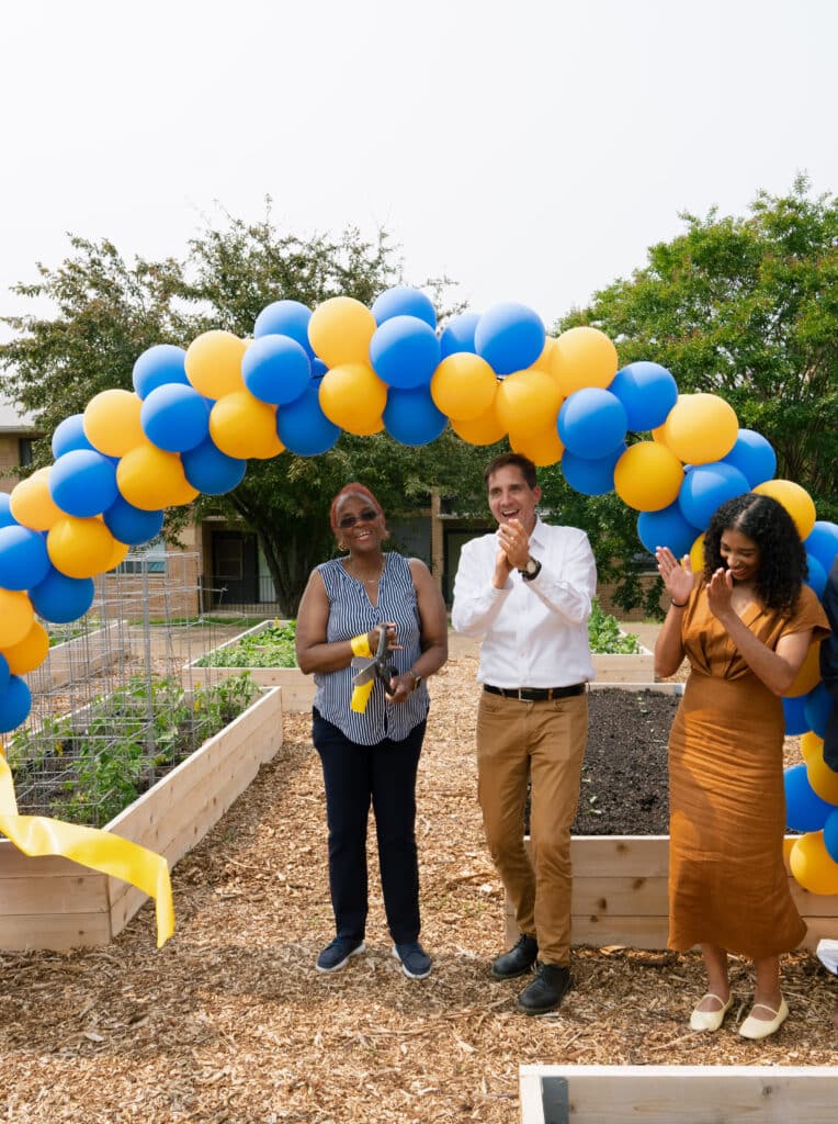 Three people stand under a blue and yellow balloon arch at a ribbon-cutting ceremony in a garden; one person holds scissors while the others clap.