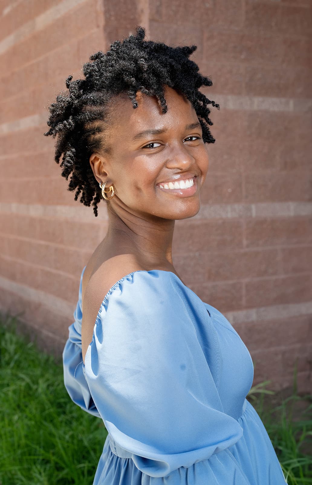 A woman in a light blue off-the-shoulder dress stands outdoors near a brick wall, smiling at the camera, embodying the spirit of building bridges and fostering connections for a brighter tomorrow.