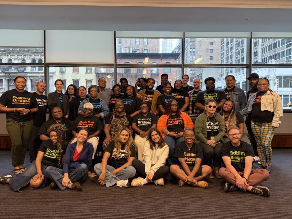 A large group of people pose for a photo indoors, many wearing shirts that say "Building Bridges." The background shows windows with a cityscape view. Discover Building Bridges Across The River - Join Us as a Volunteer!.